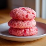 A stack of three bright pink, sugar-coated Maraschino cherry sugar cookies on a light plate.