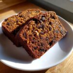 Two thick slices of moist date nut bread, rich brown in color and packed with dark raisins and nuts, resting on a white plate.