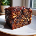 Close-up of a rich, dark slice of moist fruit cake packed with dried fruits and walnuts, sitting on a white plate.