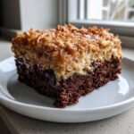 Close-up of a moist slice of german chocolate poke cake showing a rich chocolate base and thick coconut-pecan topping.