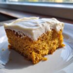 A close-up, sunlit slice of moist pumpkin poke cake topped with thick white cream cheese frosting on a white plate.