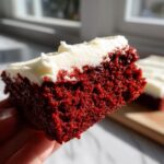 A hand holding a thick, moist square of bright red velvet brownies topped with white cream cheese frosting.