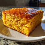 A close-up, sunlit slice of vibrant orange sweet potato cornbread drizzled with a shiny glaze on a white plate.