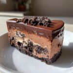 A rich slice of no-bake chocolate cheesecake featuring a dark crust, mousse layer, and glossy chocolate ganache topping.