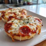 Two delicious pepperoni english muffin pizza halves topped with melted mozzarella cheese and dried herbs on a white plate.