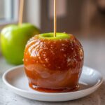 Close-up of a shiny, sticky caramel apple on a white plate, with a second green apple visible in the background.