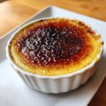 Close-up of a single serving of Creme Brulee in a white ramekin, featuring a deeply caramelized, crackling sugar crust.