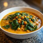 A close-up of a white bowl filled with vibrant orange pumpkin curry featuring chickpeas and wilted spinach.