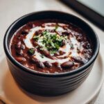 Close-up of a hearty bowl of dark, rich black bean soup topped with a swirl of sour cream and fresh cilantro.