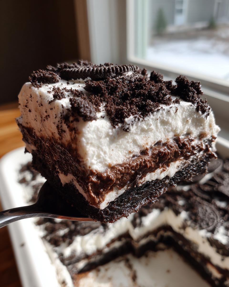 A close-up of a square serving of Oreo dirt cake being lifted on a fork, showing layers of crust, pudding, and whipped topping.