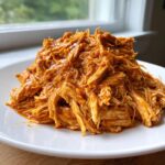 A mound of saucy, shredded buffalo chicken piled high on a white plate, ready to serve.