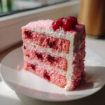 A tall slice of pink layered cherry cake with white cream frosting and whole cherries on top.