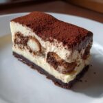 A perfect slice of tiramisu cheesecake featuring layers of cream, coffee-soaked ladyfingers, and a dark crust, topped with cocoa powder.