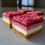 Close-up of layered strawberry crunch cheesecake bars showing graham cracker crust, white cheesecake, pink filling, and crunchy topping.