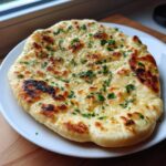 A freshly cooked, fluffy naan bread brushed with butter and topped with chopped green herbs, resting on a white plate.