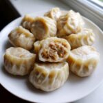 A plate of freshly steamed Traditional Chinese Dumplings, one cut open to show the savory meat filling inside.