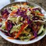 A close-up of a fresh cabbage salad featuring shredded green and purple cabbage, carrots, and cucumber slices.