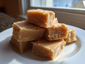 A small stack of creamy, light brown maple fudge squares sitting on a white plate near a window.
