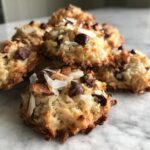 A close-up of several golden-brown almond joy cookies topped with chocolate chips, coconut flakes, and sliced almonds.