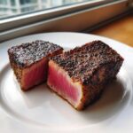 Two thick slices of perfectly seared tuna steak, showing a bright pink rare center and a dark, peppery crust.