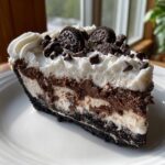 A decadent slice of 7-layer ice cream pie featuring layers of chocolate and cream, topped with whipped cream, mini chocolate chips, and Oreo cookies.