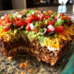Close-up of a 7 layer dip with a scoop removed, showing layers of meat, cream cheese, guacamole, cheese, tomatoes, and olives.
