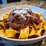 A close-up of wide pappardelle pasta topped generously with shredded beef ragu and grated Parmesan cheese.