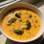 A close-up shot of a creamy, orange-colored bowl of broccoli cheddar soup topped with fresh broccoli florets.