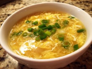 A close-up of a white bowl filled with hot, yellow egg drop soup, garnished with fresh green scallions.