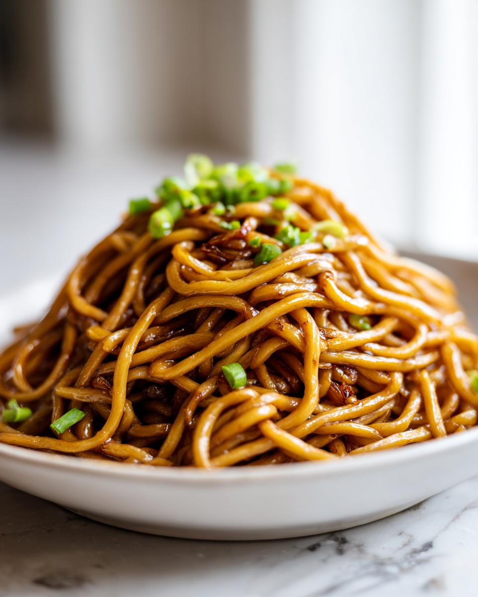 A close-up shot of a generous serving of glossy, brown garlic noodles topped with chopped green onions.