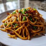 A close-up of a serving of saucy, thick garlic noodles topped with green onions and crispy fried bits.