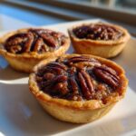 Three freshly baked mini pecan pies with glossy pecan toppings sitting on a white plate.