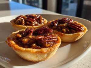 Close-up of three glossy, golden mini pecan pies with whole pecans on top, sitting on a white plate.