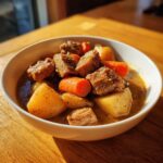 A close-up of a white bowl filled with rich, savory pork stew chunks, potatoes, and bright orange carrots.