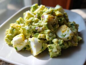 A close-up, bright photo of creamy avocado egg salad mixed with chopped hard-boiled eggs and herbs on a white plate.
