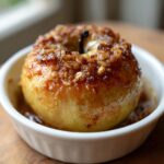 A single, glistening baked apple topped with a brown sugar and oat crumble, served in a white ramekin.