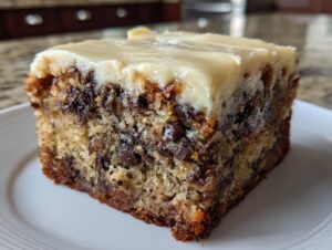 Close-up of a moist square slice of banana bread brownies loaded with chocolate chips and topped with thick cream cheese frosting.