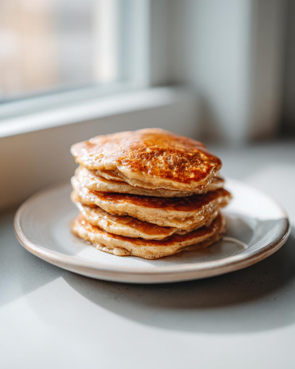 A close-up stack of five golden brown banana oatmeal pancakes drizzled with syrup, sitting on a light plate.