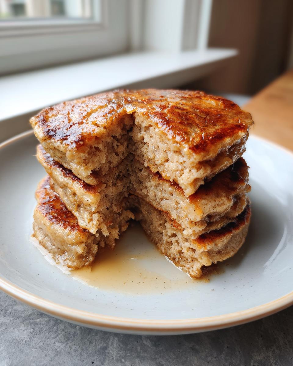 A stack of thick banana oatmeal pancakes with a piece cut out, drizzled with syrup.