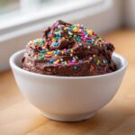 A close-up of rich, chocolate brownie batter dip topped generously with colorful rainbow sprinkles in a white bowl.
