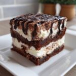 A square slice of layered brownie refrigerator cake with cream filling and chocolate drizzle.