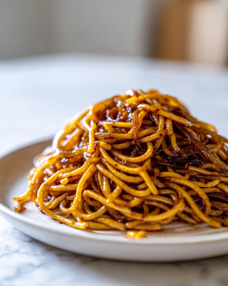A close-up of a mound of glossy caramelized onion pasta coated in a rich, dark sauce on a light plate.