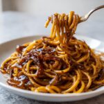 A fork lifting strands of glossy caramelized onion pasta coated in a rich, dark sauce.