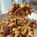 A fork lifts a cheesy bite of macaroni noodles topped with caramelized onions from a bowl of french onion pasta.