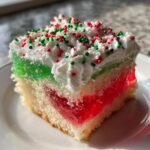 A festive slice of christmas poke cake showing layers of white cake, red and green jello, whipped topping, and sprinkles.