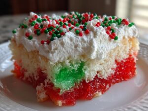 A close-up slice of christmas poke cake showing layers of white cake, red and green jello, whipped topping, and sprinkles.