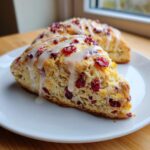 A close-up of a freshly baked cranberry orange scone topped with white vanilla glaze, showing dried cranberries inside.