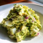 A mound of creamy avocado egg salad featuring chunks of white egg, green avocado, and flecks of red onion on a white dish.