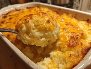 A spoonful of creamy baked mashed potatoes topped with melted, browned cheddar cheese being lifted from the casserole dish.