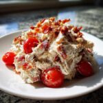 A serving of creamy blt chicken salad generously topped with crispy bacon crumbles and whole cherry tomatoes on a white plate.